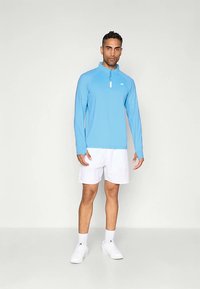 Šviesiai mėlynas ilgomis rankovėmis sportinis viršus su ketvirčio užtrauktuku, baltos šortai ir balti sportbačiai. Lygus audinys, aptemptas dizainas ir minimalus prekės ženklinimas.