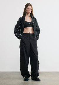 Femme portant une veste noire raccourcie, un brassière de sport noire et un pantalon cargo noir ample, les mains dans les poches, debout contre un mur blanc uni.