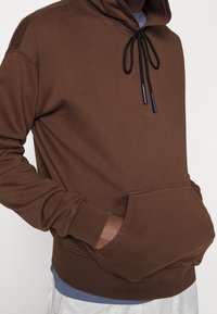 Felpa marrone con cappuccio e lacci, tasca frontale e polsini a coste. Il materiale appare morbido e testurizzato, con una vestibilità comoda. Accenti blu sottili.