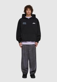 Felpa nera con grafiche blu e bianche, tasca a marsupio, abbinata a pantaloni grigi a vestibilità comoda. Il modello indossa sneakers nere.