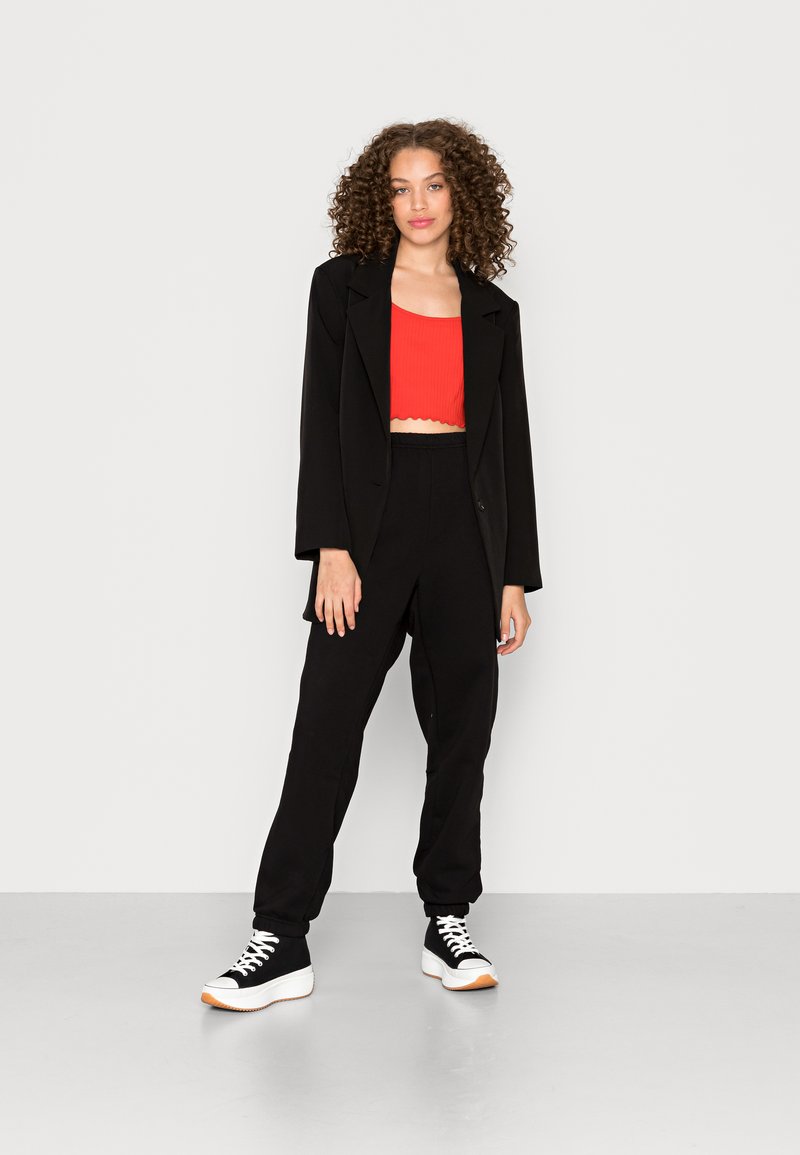Blazer oversized preto, top cropped vermelho com nervuras, calças pretas ajustadas e ténis pretos com solas brancas; cabelo encaracolado e fundo neutro.