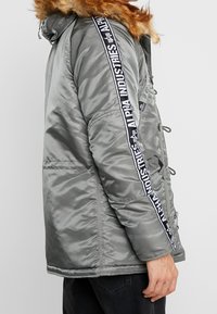 Grå vadderad jacka med huva i konstpäls, med en svart och vit sidologo-stripe, texturerat tyg och flera fickor.