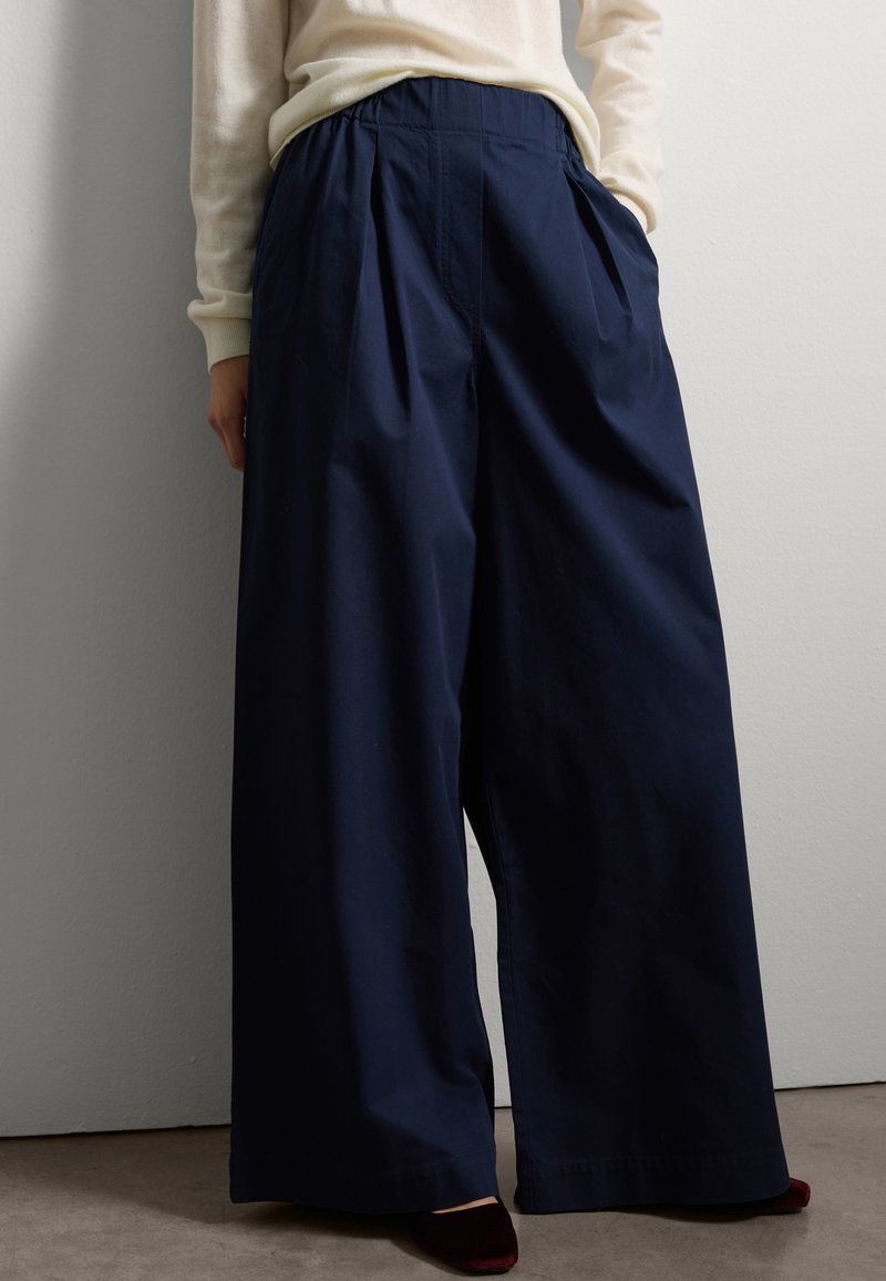 Pantalon large marine en coton, avec une taille froncée, des plis et une texture lisse. Porté avec un haut de couleur claire et des chaussures.