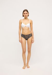Brasile bianca con spalline sottili abbinata a mutandine in pizzo scuro, con intricati motivi floreali e bordi ondulati. La modella è in piedi a piedi nudi.