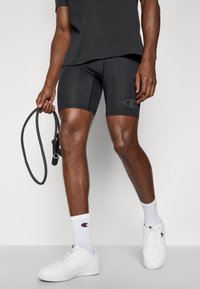 Zwarte compressiekorte broeken met een logo, gecombineerd met witte sport sneakers en sokken met een logo. Het model houdt fitnessapparatuur vast.