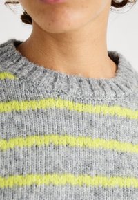 Harmaa neulepusero, jossa vaakasuorat neonkeltaiset raidat. Paksu resorikaulus ja pehmeä rakenne. Tarkennus kankaaseen ja värikontrastiin.