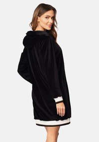 Černé sametové šaty s kapucí a volným střihem, bílými ribbovanými manžetami a jemnou texturou. Obsahují kapuci a zakřivený lem.