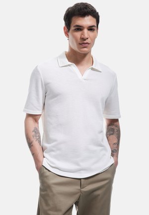 Polo blanco de manga corta con un patrón texturizado, cuello en V y ribete del cuello en contraste, combinado con pantalones beige.