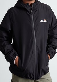 Persona che indossa una giacca a zip con cappuccio nera con il logo Ellessee sul petto, mani nelle tasche, e pantaloni beige visibili sotto.