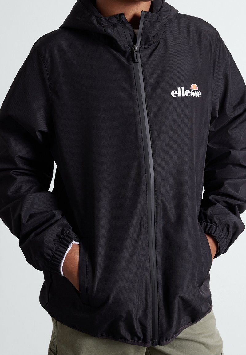 Persona che indossa una giacca a zip con cappuccio nera con il logo Ellessee sul petto, mani nelle tasche, e pantaloni beige visibili sotto.