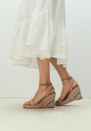 Nohy v béžových pletených klinových sandáloch s členkovými remienkami a bielych šatách s textúrovaným, vrstveným lemom.