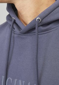Mikina s kapucňou v tmavo sivej tkanine, s prednou kapsou, šnúrkami na kapucni a diskrétnym logom pri okraji. Mäkkučká textúra s uvoľneným strihom.