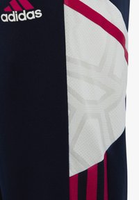Pantalones deportivos de color navy con acentos en blanco y rojo, textura de malla sintética, logo icónico y patrón geométrico en el panel lateral.