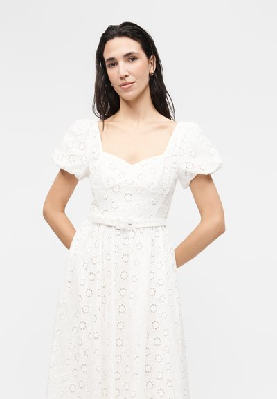 Γυναίκα με μακριά σκούρα μαλλιά, που φοράει λευκό φόρεμα με διάτρητο σχέδιο, φουσκωτά μανίκια και εφαρμοστή μέση, στέκεται με τα χέρια πίσω από την πλάτη της.