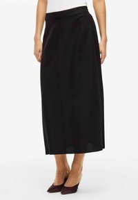 Jupe longue noire en tissu doux, présentant une silhouette droite et une taille. Portée avec des chaussures bordeaux à bout pointu.