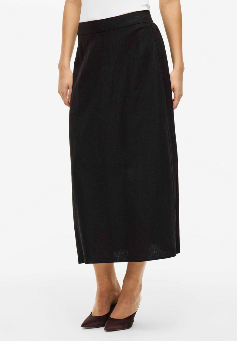 Jupe longue noire en tissu doux, présentant une silhouette droite et une taille. Portée avec des chaussures bordeaux à bout pointu.