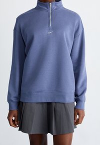 Sweatshirt i ljusblått tyg, med hög krage, halvvisslad framtill och en liten silverlogotyp på bröstet.