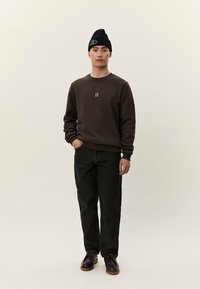 Hombre con gorro negro, sudadera marrón oscuro con el logo "II", vaqueros negros y zapatos marrón oscuro, de pie frente a un fondo liso.