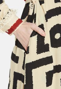 Beige mekko, jossa on rohkeita mustia abstrakteja kuvioita, sivutasku ja fringejä hihoissa punaisella yksityiskohdalla. Valmistettu kevyestä kankaasta.
