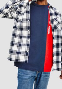 Camicia di flanella a quadri in navy, bianco e grigio sopra una maglietta blu e rossa a blocchi di colore con la scritta "TOMMY JEANS" in lettere bianche. Jeans di denim.