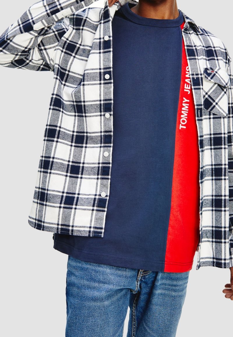 Camicia di flanella a quadri in navy, bianco e grigio sopra una maglietta blu e rossa a blocchi di colore con la scritta "TOMMY JEANS" in lettere bianche. Jeans di denim.