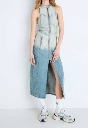 Femme portant une robe en jean dégradé bleu sans manches avec fermeture éclair devant et fente latérale, chaussettes blanches et baskets Nike argentées.