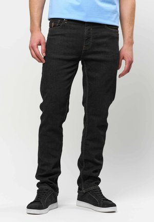 Homme portant un jean noir slim et des baskets noires avec des semelles blanches, debout devant un fond clair uni.