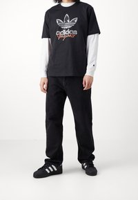 Černé bavlněné tričko s grafikou loga Adidas. Nosené přes bílé tričko s dlouhým rukávem. Slučuje se s volnými černými džíny a černými teniskami s bílými pruhy.