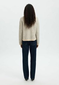 Personne avec de longs cheveux bouclés portant un pull en maille beige, un jean bleu foncé et des chaussures à talons noires, debout de dos sur un fond blanc.