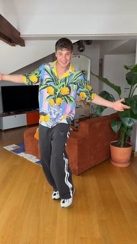 Jeune homme souriant les bras écartés, portant une chemise colorée à motif citron, un pantalon noir et des baskets blanches dans un salon.