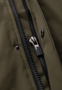 Nahaufnahme eines schwarzen Reißverschlusses auf olivgrünem Stoff mit einem Metallreißverschluss-Zipper und einem kleinen Metall-Druckknopf in der Nähe.
