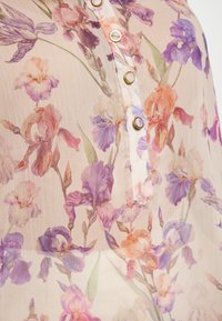 Durchsichtige Bluse in hellem Beige mit buntem Iris-Blumenmuster. Verfügt über eine Knopfleiste mit goldfarbenen Knöpfen und zarter Textur.