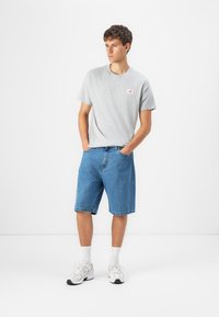Grå t-skjorte med en liten logo og blå denimshorts. Modellen har på seg hvite sneakers og hvite sokker, står med hendene i lommene.