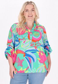 Blusa colorata a maniche corte con un vivace motivo floreale in verde, rosa, blu e giallo. Presenta un collo a bottoni e volant.