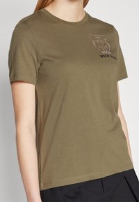 Camiseta de algodón color verde oliva con mangas cortas, escote redondo y un gráfico texturizado que presenta un oso y el texto "WILD BOAR" en strass.