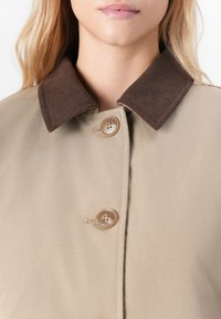 Manteau beige avec un col en velours côtelé marron, comportant deux boutons et une texture lisse. Accent sur les détails du col et des boutons.