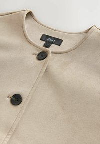 Veste en tissu beige et doux avec un col arrondi, dotée de deux boutons noirs et d'une étiquette noire avec "NEXT" au niveau du col.