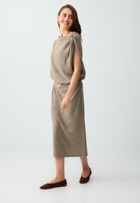 Femme portant un haut sans manches taupe froncé et une jupe midi assortie, souriante et regardant sur le côté, debout sur fond blanc.