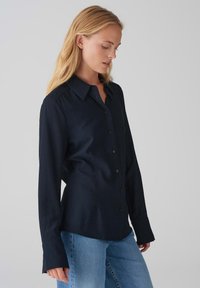 Camicia blu navy con bottoni in un tessuto morbido, colletto, maniche lunghe e una vestibilità comoda, abbinata a jeans azzurri.