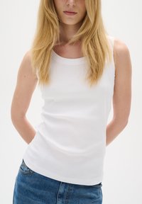 Femme aux longs cheveux blonds portant un haut blanc sans manches côtelé et un jean bleu en denim, debout les mains dans le dos.