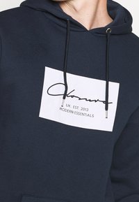 Felpa con cappuccio blu navy con una patch rettangolare bianca che presenta una firma. Cappuccio con cordino e punte in metallo, realizzato in tessuto morbido.