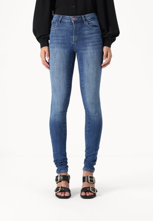 Fitted blue jeans in a medium wash, featuring five pockets, a button closure, and a smooth denim texture. Paired with black sandals.