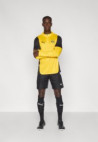 Στολή ποδοσφαίρου που περιλαμβάνει κίτρινη και μαύρη μακρυμάνικη μπλούζα με φερμουάρ, μαύρο σορτς, μαύρες κάλτσες και μαύρα παπούτσια με αγκράφες, με λεπτομέρειες λογότυπων.