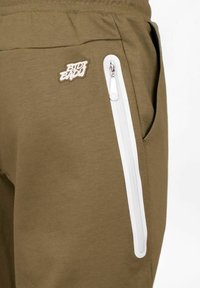 Olivgrüne Jogginghose mit einer weißen Zipper-Tasche, aus einem strukturierten Stoff und einem kleinen gestickten "Bidi Badu"-Logo am Oberschenkel.