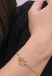Bracciale in oro con design a cuori intrecciate. Il cuore presenta piccole pietre trasparenti lungo il bordo. Texture della catena liscia. Elegante e delicato.