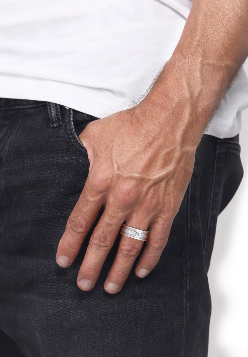 Sølvring med rosaguldbånd med tre horisontale striber og en lille central ædelsten, båret på en hånd, der hviler i en lomme.