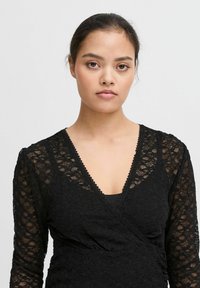 Top avvolgente in pizzo nero con maniche lunghe, caratterizzato da una profonda scollatura a V, bordi smerlati e un motivo floreale testurizzato. Strato interno liscio visibile.