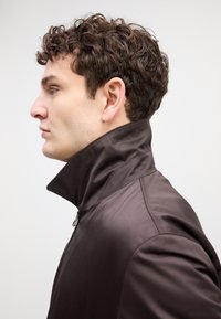 Veste marron à col haut avec une texture lisse. Fermeture éclair sur le devant et design épuré, mettant en valeur une silhouette moderne vue de côté.