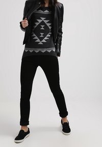 Veste en cuir noire sur un pull gris à motif géométrique, associée à un pantalon slim noir et des chaussures slip-on noires.