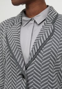 Blazer en chevrons gris avec un col contrasté, fermeture par bouton et coutures détaillées. Porté sur une chemise à collier gris clair.